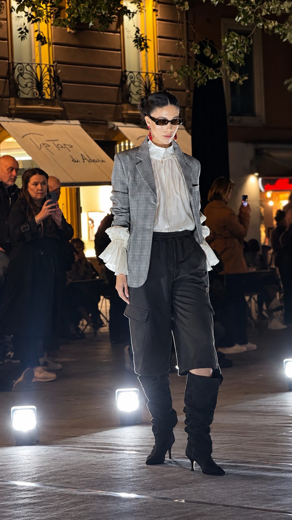 Una modella con blazer grigio, camicia bianca con polsini a balze, bermuda neri e stivali alti, mentre il pubblico la riprende con il telefono