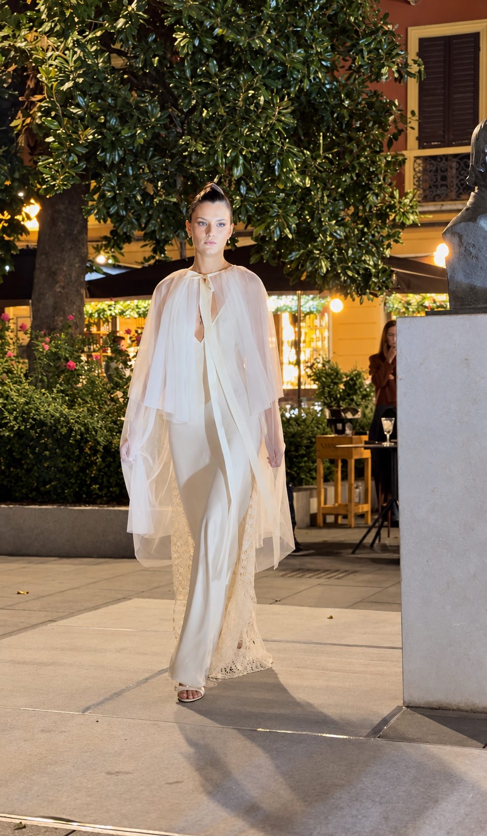 Una modella in total white, con un capo lungo e leggero sopra l'abito, sfila sotto gli alberi illuminati